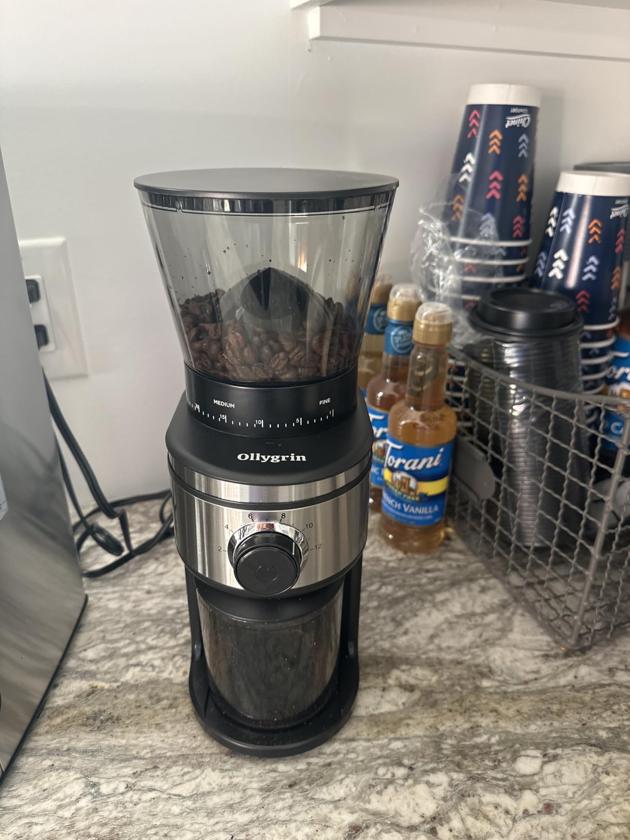 Ollygrin conical burr grinder with beans in the hopper on a home coffee station beside syrup bottles and paper cups